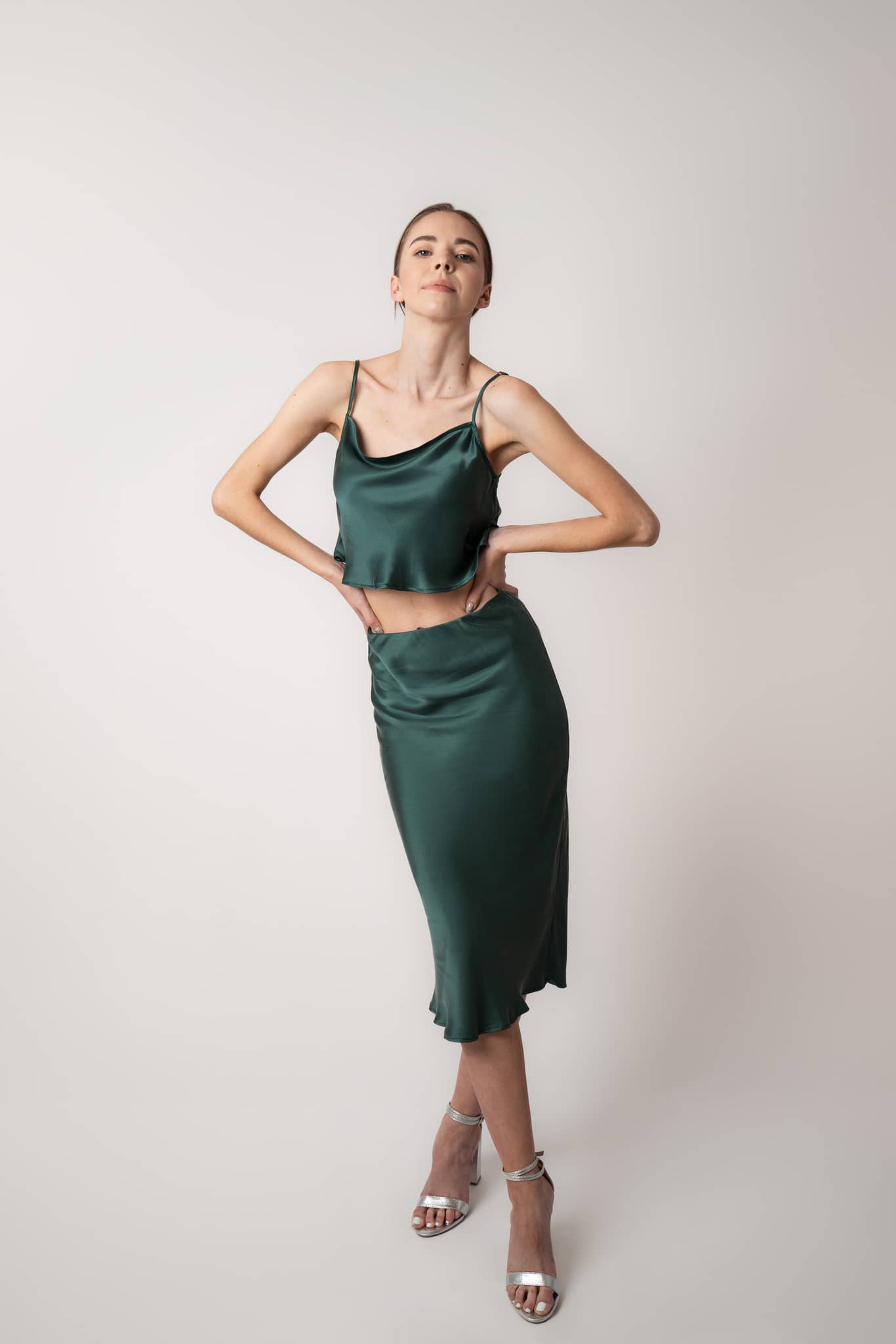 Woman sitting on couch wearing Silk Closet cowl neck sandwash silk green crop top and skirt in studio. Party outfit.